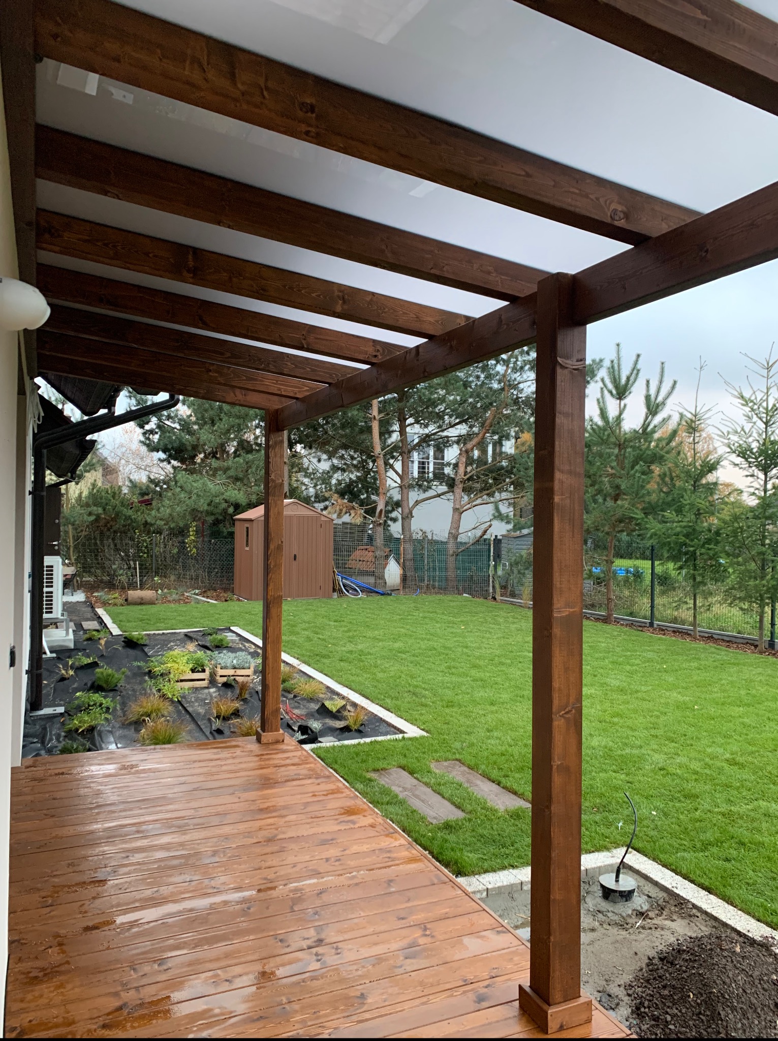 Zadaszony taras z drewna z widokiem na zadbany ogród. Elementy drewniane w ciepłym odcieniu brązu, zadaszenie z białych paneli. W tle zielony trawnik i drzewa.