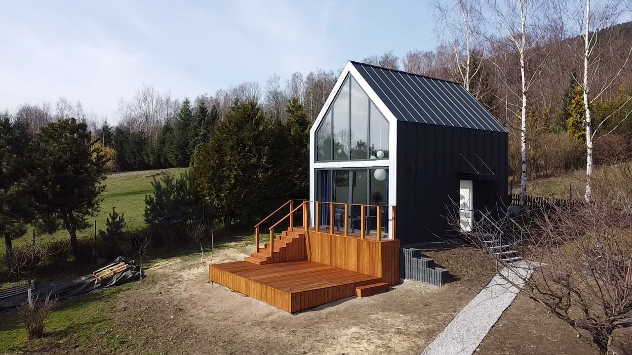 Nowoczesny dom z drewnianym tarasem w Sobótce. Widok z góry ukazuje schody i balustrady. Tło stanowią drzewa i wzgórza, tworząc malowniczy krajobraz.