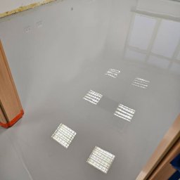 VISIONFLOOR MICHAŁ GRYZIO - Jasnoszara, gładka posadzka z widocznymi odbiciami lamp sufitowych. Przy ścianach listwy przypodłogowe, widoczne gniazdka elektryczne. Fragment ościeżnicy drzwi.