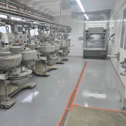 VISIONFLOOR MICHAŁ GRYZIO - Szara, gładka posadzka w hali produkcyjnej z rzędem mieszalników przemysłowych. Widoczne rury pod sufitem i białe kafelki na ścianach. Pomarańczowe akcenty na podłodze.