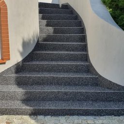 VISIONFLOOR MICHAŁ GRYZIO - Schody zewnętrzne z szarej, drobnoziarnistej nawierzchni, z białymi listwami na krawędziach stopni. Obok okno z czerwoną cegłą i zielony krzew.