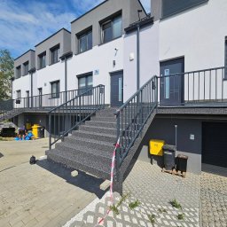 VISIONFLOOR MICHAŁ GRYZIO - Zewnętrzne schody wejściowe do szeregowca wykończone szarą, kamyczkową posadzką. Widoczne barierki, skrzynki na listy i fragment chodnika z kostki brukowej.
