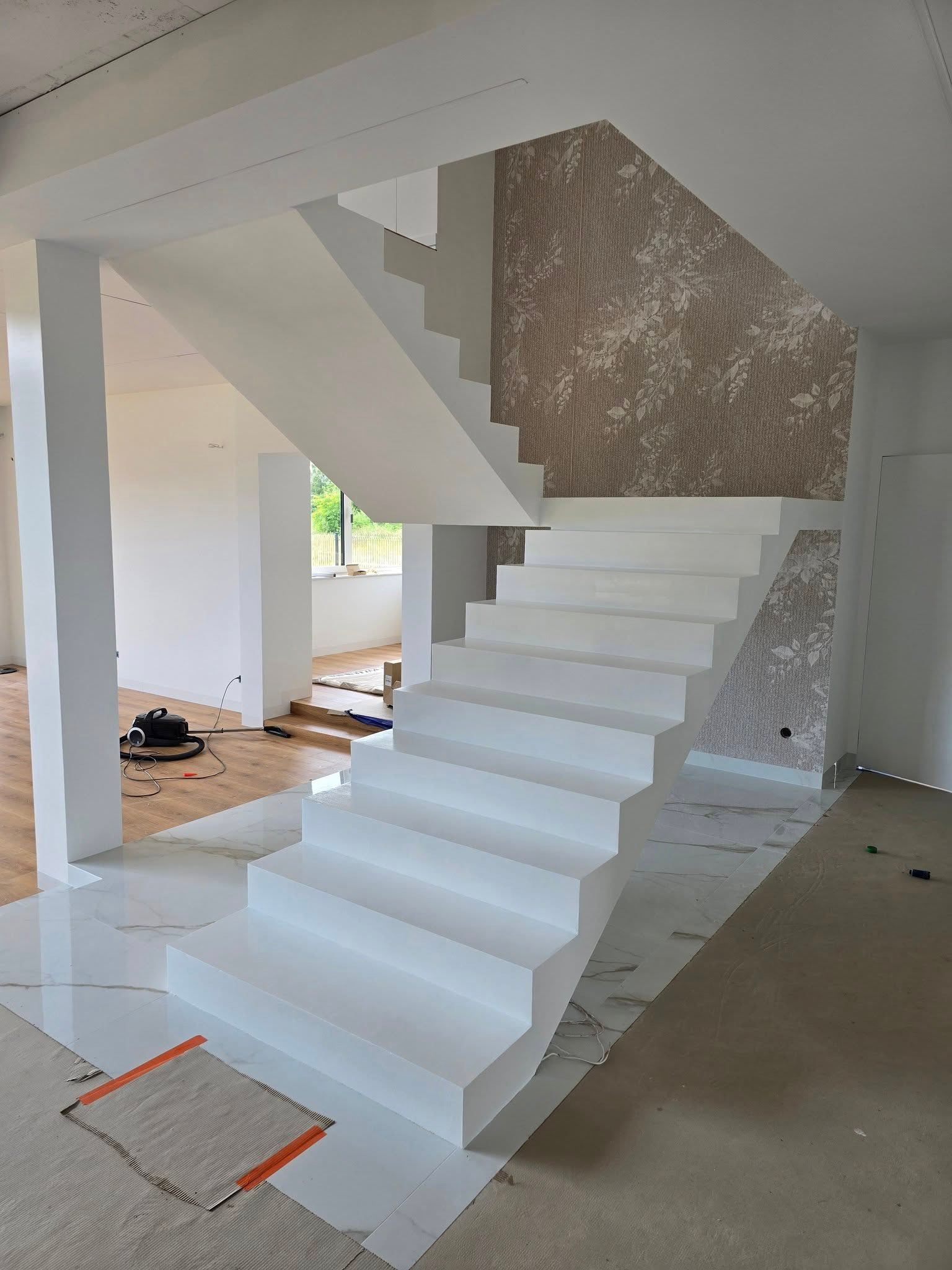 Nowoczesne, białe schody w jasnym wnętrzu z marmurową posadzką i wzorzystą tapetą. Widoczny odkurzacz i zabezpieczenia podłogi, sugerujące prace wykończeniowe.
