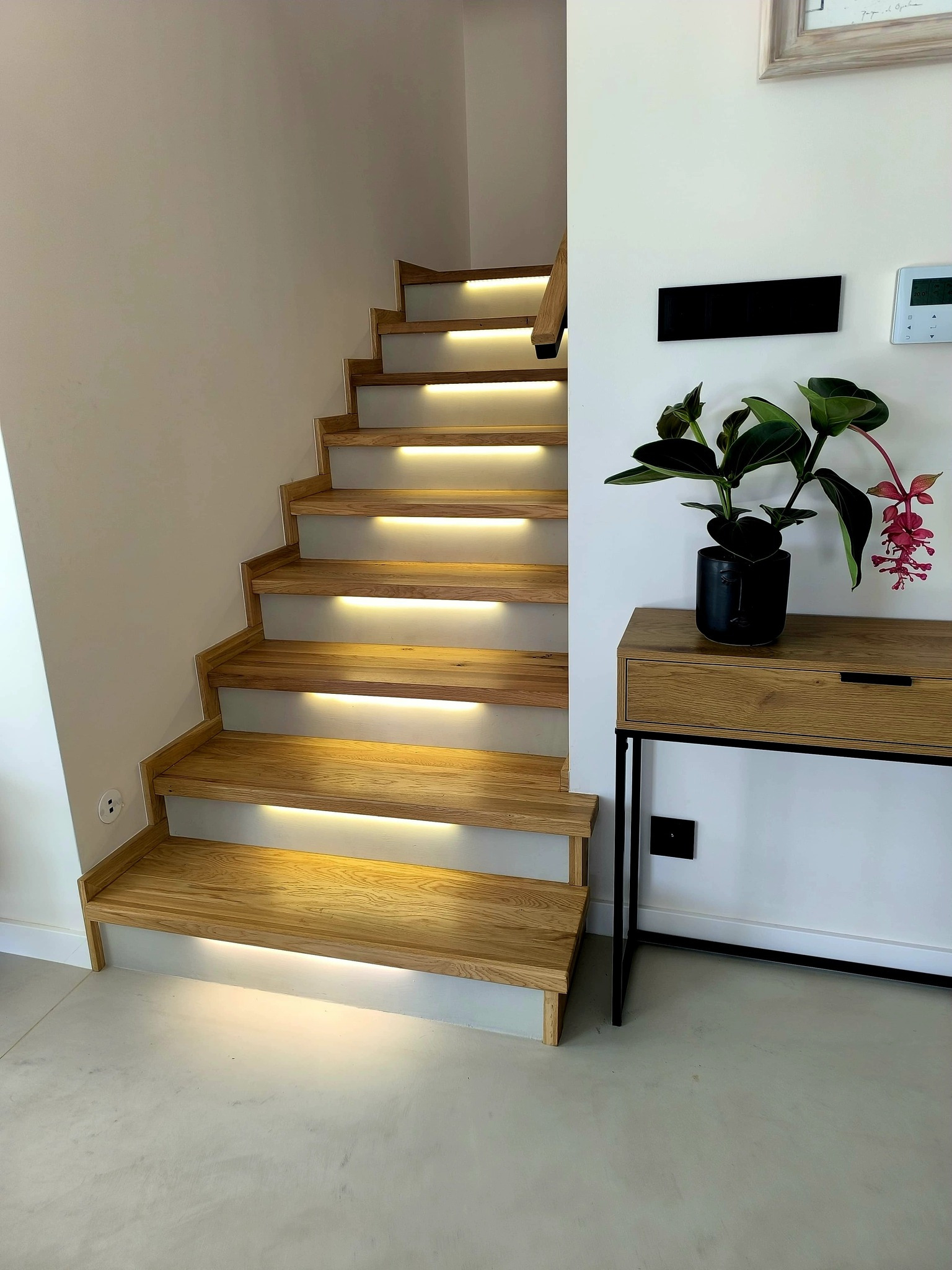 Drewniane schody z podświetleniem LED na białej ścianie, obok konsola z rośliną. Minimalistyczny design, jasne wnętrze, gładka, jasna posadzka.