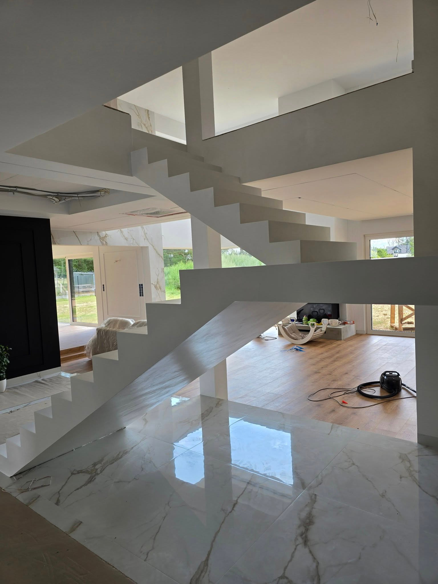 Nowoczesne, białe schody z mikrocementu w przestronnym wnętrzu. Błyszcząca posadzka z marmurowym wzorem odbija światło. Widoczny odkurzacz i fragment salonu.