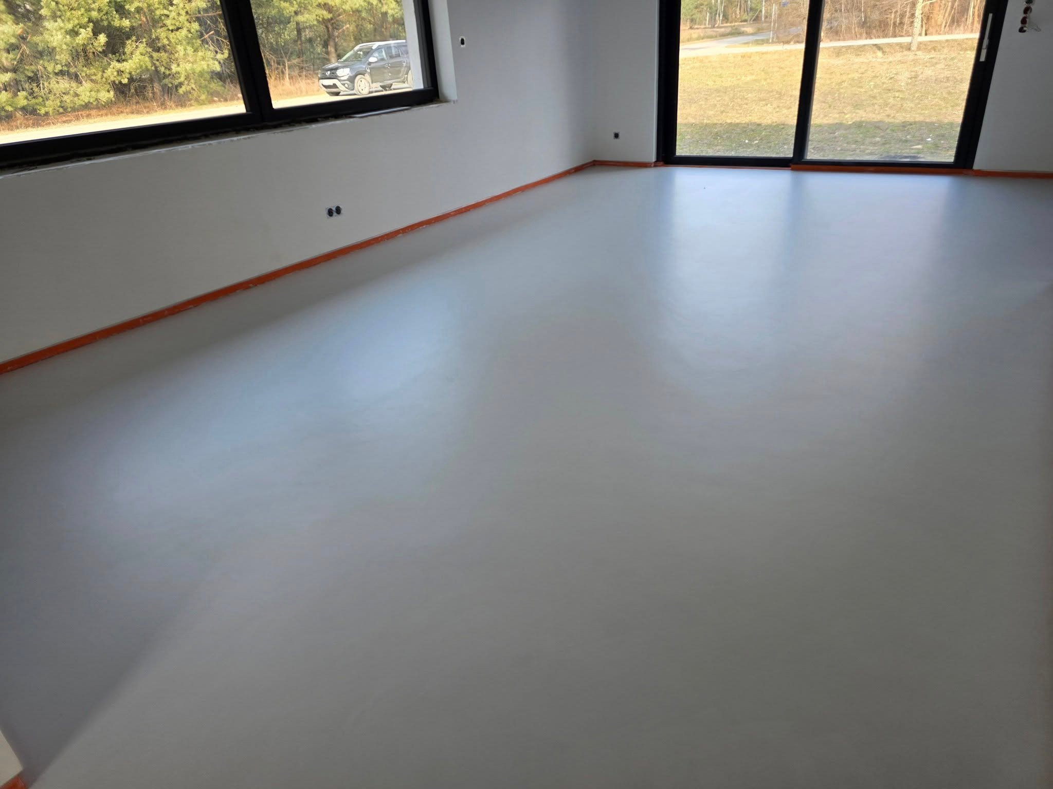 Jasnoszara, gładka posadzka w nowym wnętrzu z dużymi oknami. Widoczne listwy przypodłogowe i fragment krajobrazu za oknem. Minimalistyczny design.