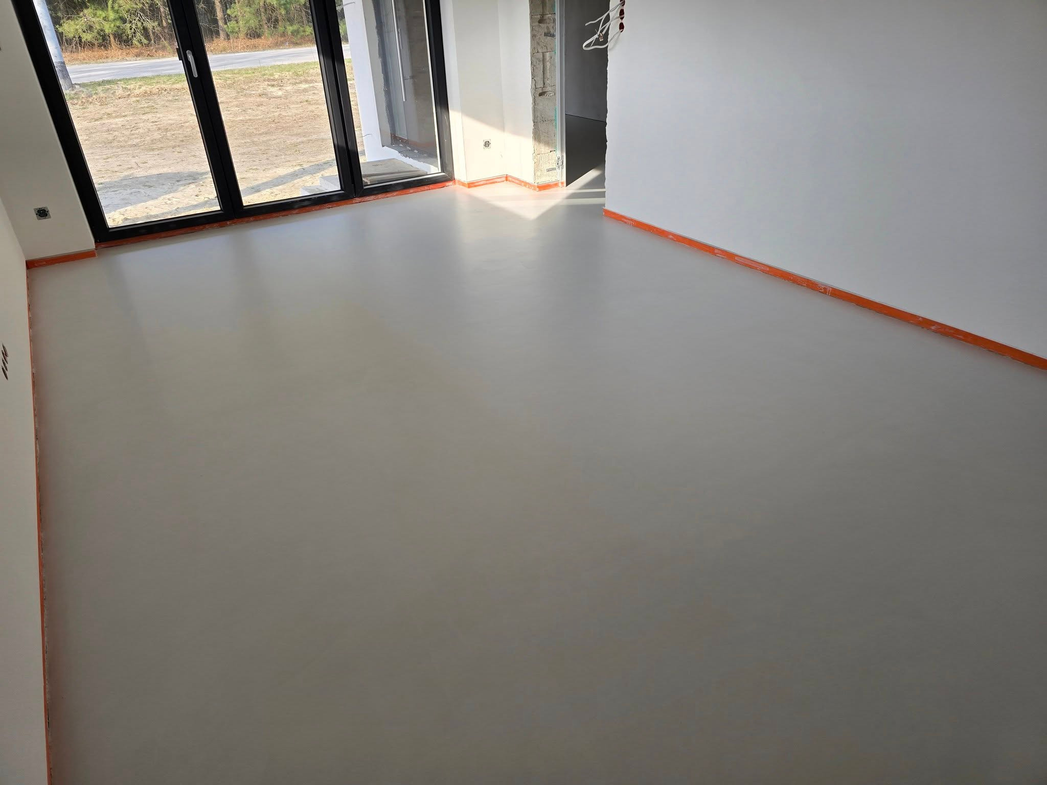 Jasnoszara, gładka posadzka w nowym wnętrzu z dużym oknem balkonowym i pomarańczowymi listwami przypodłogowymi. Widok na zewnątrz i do sąsiedniego pomieszczenia.