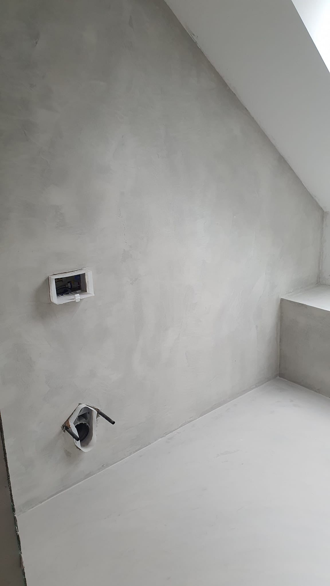 Jasnoszara, gładka powierzchnia ściany i podłogi w pomieszczeniu z widocznymi otworami na instalacje elektryczne. Minimalistyczne wykończenie wnętrza.