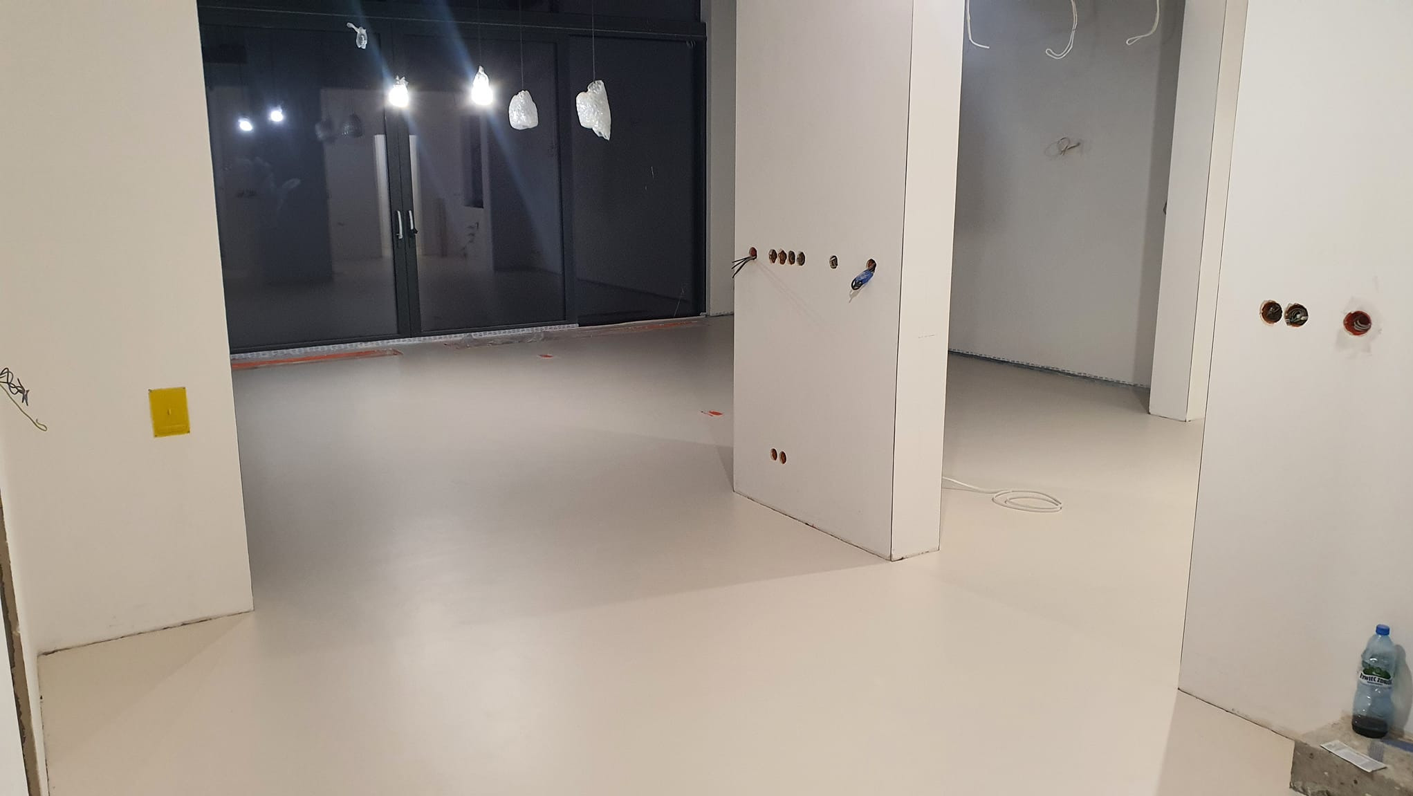 Jasna, gładka posadzka w nowym wnętrzu z widocznymi instalacjami elektrycznymi w ścianach. Minimalistyczny design, duże okna z czarnymi ramami.