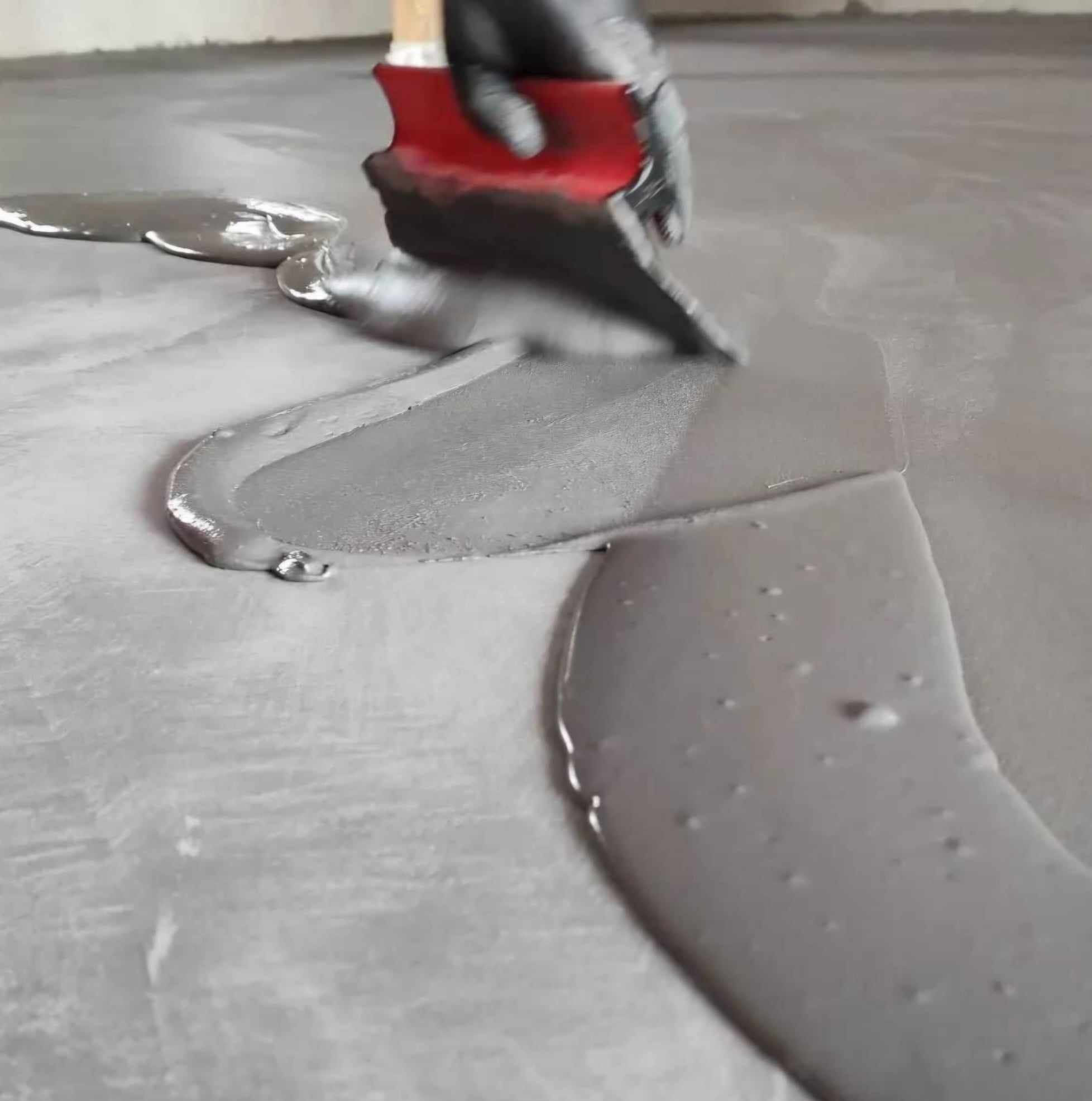 Aplikacja szarej masy na podłodze za pomocą czerwonej szpachelki, widoczne ślady pracy i gładka powierzchnia. Ręka w czarnej rękawicy. Detal wykończenia.