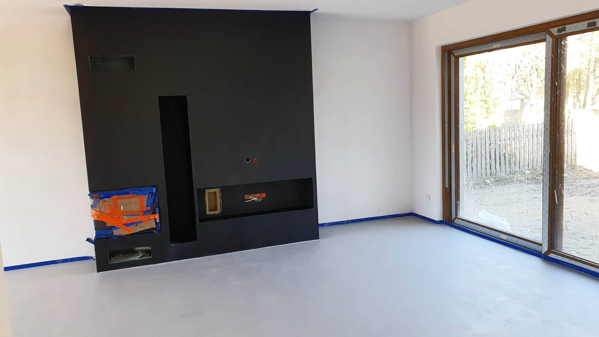 Jasnoszara posadzka w surowym wnętrzu z czarną ścianą kominkową. Okno z widokiem na zewnątrz. Prace wykończeniowe w toku, zabezpieczenia taśmą malarską.