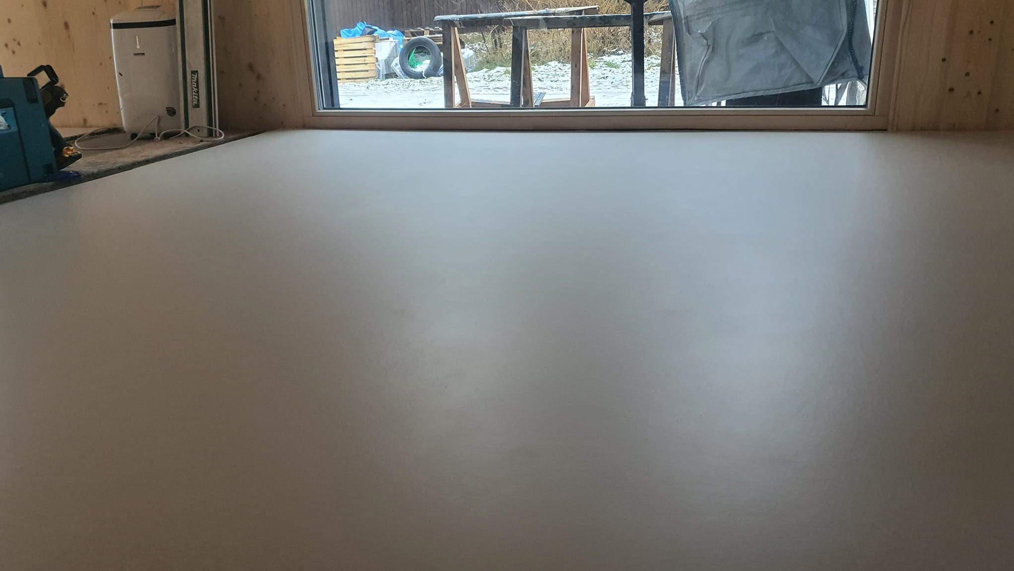 Gładka, jasnoszara posadzka w pomieszczeniu z drewnianymi ścianami i oknem. Widoczne narzędzia i sprzęt budowlany w tle. Minimalistyczny design.