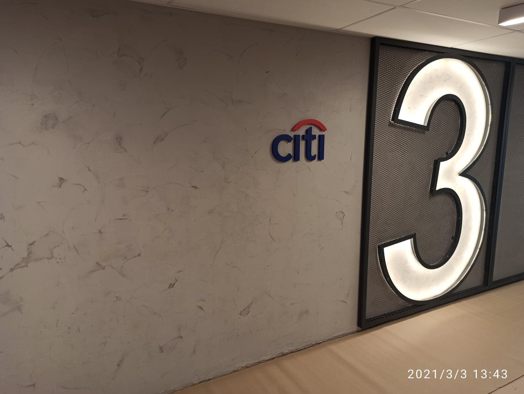 Ściana z efektem dekoracyjnym w odcieniach szarości, logo Citi i podświetlona cyfra 3 w metalowej ramie. Nowoczesne wykończenie wnętrza biurowego.