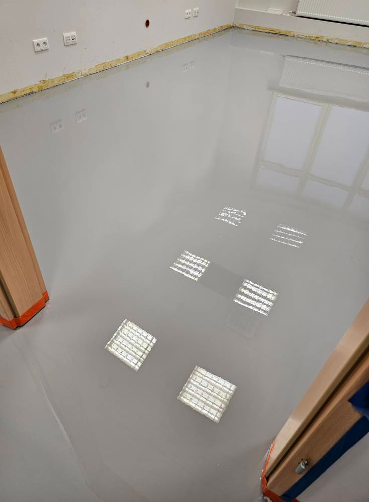 Jasnoszara, gładka posadzka z widocznymi odbiciami lamp sufitowych. Przy ścianach listwy przypodłogowe, widoczne gniazdka elektryczne. Fragment ościeżnicy drzwi.