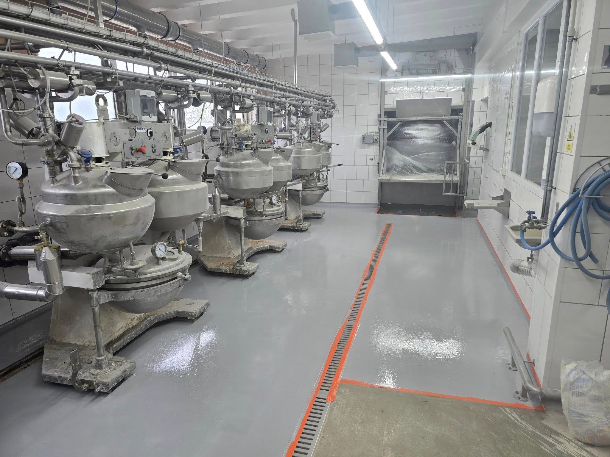 Szara, gładka posadzka w hali produkcyjnej z rzędem mieszalników przemysłowych. Widoczne rury pod sufitem i białe kafelki na ścianach. Pomarańczowe akcenty na podłodze.