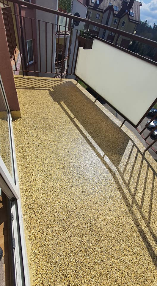 Żółta, drobnoziarnista posadzka na balkonie z brązową barierką. Widok z góry, cień barierki na podłodze. Budynki w tle, błękitne niebo.