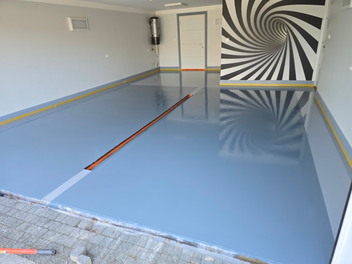 Błękitna, błyszcząca posadzka z dylatacją w garażu, z żółtą taśmą malarską przy ścianach i spiralnym wzorem na ścianie. Widoczny fragment siatki zbrojeniowej.