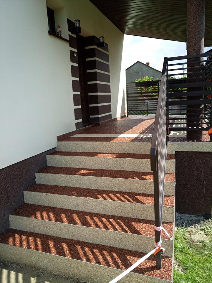 Zewnętrzne schody wejściowe z brązowo-beżową posadzką żywiczną, prowadzące do domu z kolumną i metalową barierką. Widoczne detale wykończenia elewacji.