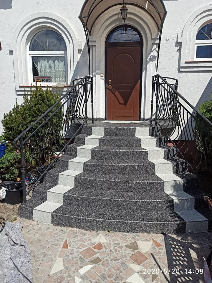 Schody wejściowe z szarą, żywiczną nawierzchnią i białymi obrzeżami, prowadzące do domu w Pasymiu. Metalowa balustrada i zadaszenie nad drzwiami.