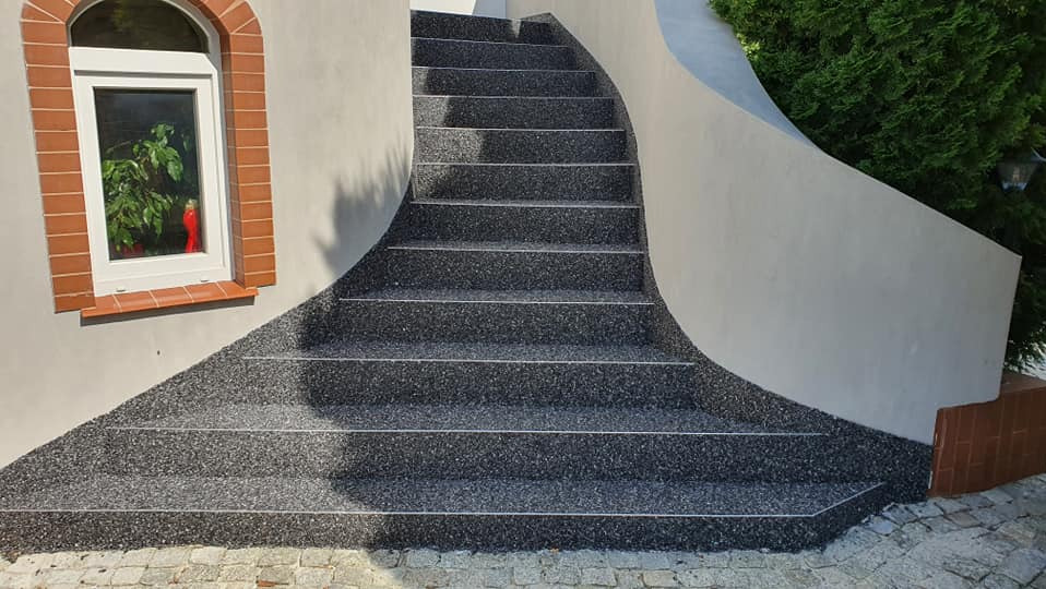 Schody zewnętrzne z szarej, drobnoziarnistej nawierzchni, z białymi listwami na krawędziach stopni. Obok okno z czerwoną cegłą i zielony krzew.