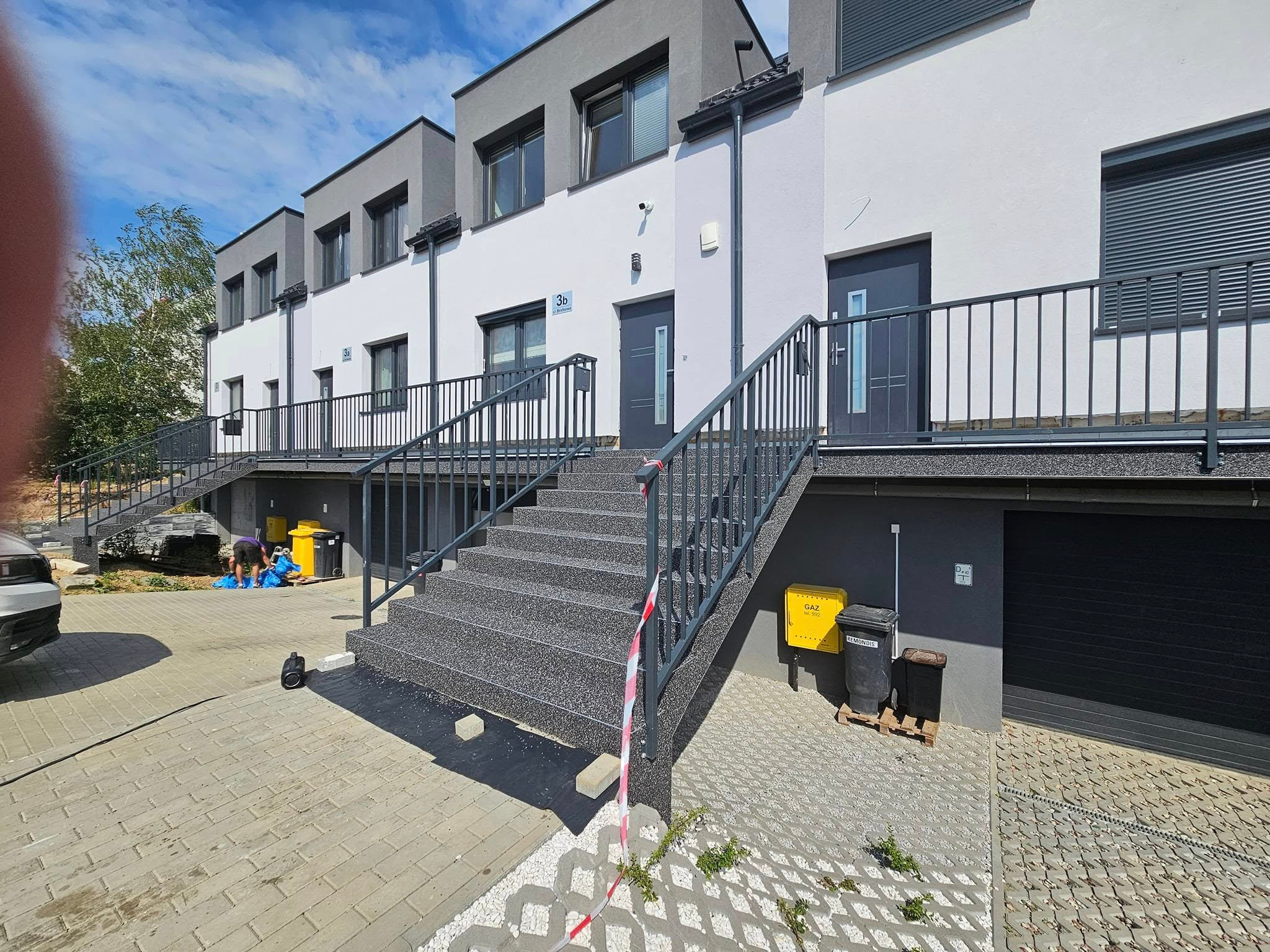 Zewnętrzne schody wejściowe do szeregowca wykończone szarą, kamyczkową posadzką. Widoczne barierki, skrzynki na listy i fragment chodnika z kostki brukowej.