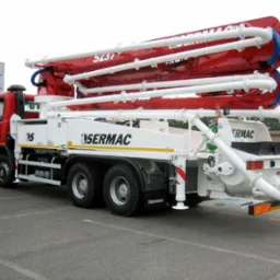 Czerwono-biała pompa do betonu marki Sermac 5Z37 zaparkowana na szarym parkingu z budynkiem w tle.