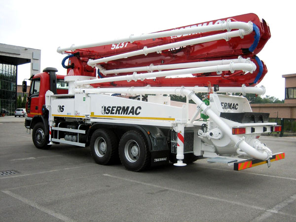 Czerwono-biała pompa do betonu marki Sermac 5Z37 zaparkowana na szarym parkingu z budynkiem w tle.