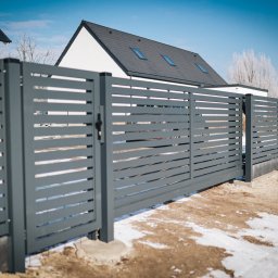LADEX OGRODZENIA RAFAŁ LADEMAN - Nowoczesna, przesuwna brama wjazdowa z poziomych paneli w kolorze grafitowym, na tle domu z ciemną dachówką i błękitnego nieba. Zimowy krajobraz z resztkami śniegu.
