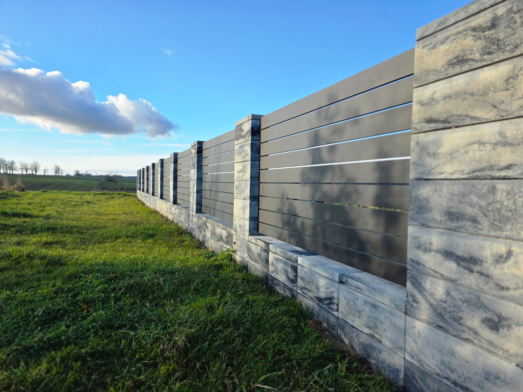 Nowoczesne ogrodzenie panelowe z szarego metalu i kamiennych słupków na tle zielonej trawy i błękitnego nieba z chmurami. Widok z perspektywy żabiej.