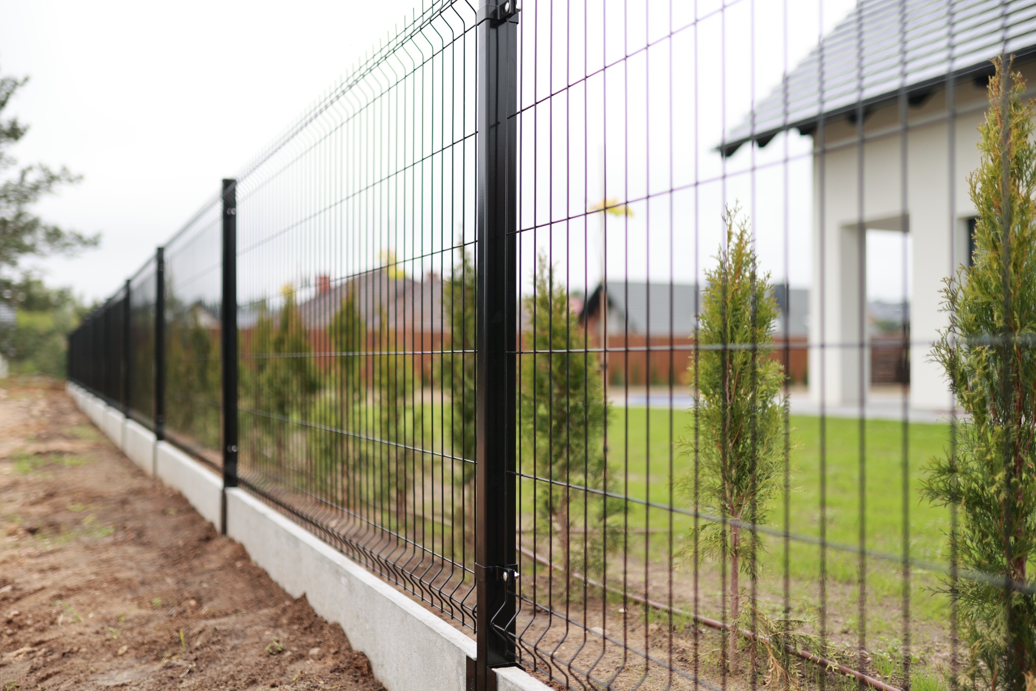 Długi, czarny panel ogrodzeniowy z podmurówką betonową, widoczny z boku, na tle trawnika z tujami i domu z szarą dachówką. Ostry kadr na ogrodzenie.