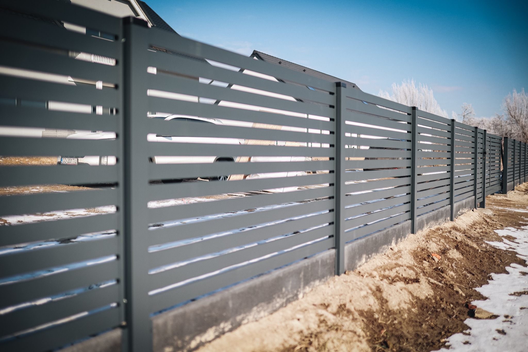 Nowoczesne, poziome ogrodzenie panelowe w kolorze grafitowym, z widocznym samochodem i ośnieżonym drzewem w tle. Ujęcie z bliska, podkreślające detale.
