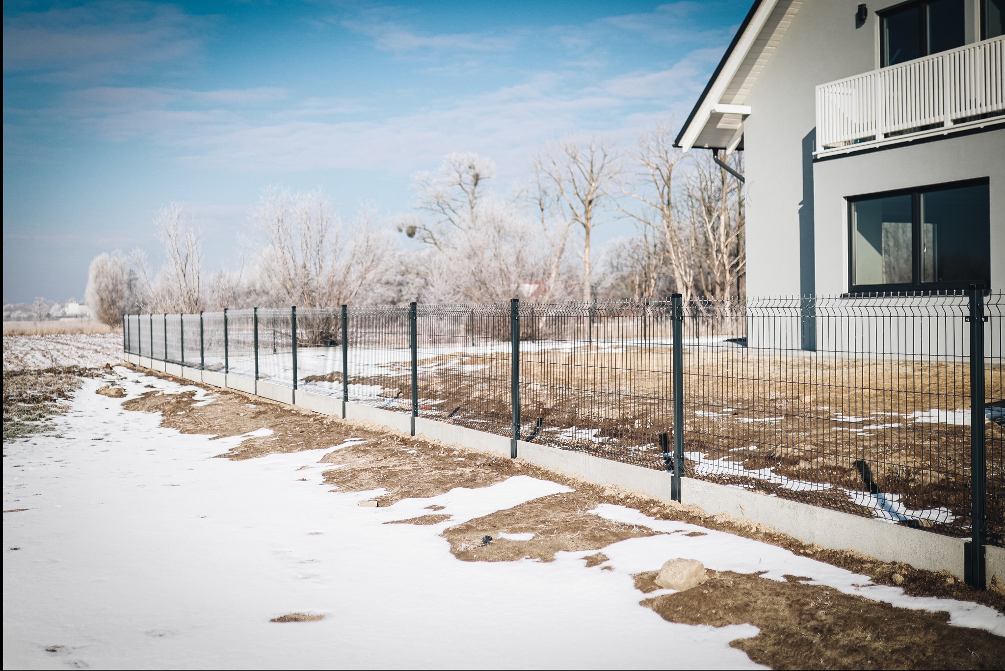 Panelowe ogrodzenie w zimowej scenerii, z widocznym fragmentem nowoczesnego domu w tle. Śnieg podkreśla kontrast z ciemną zielenią paneli.