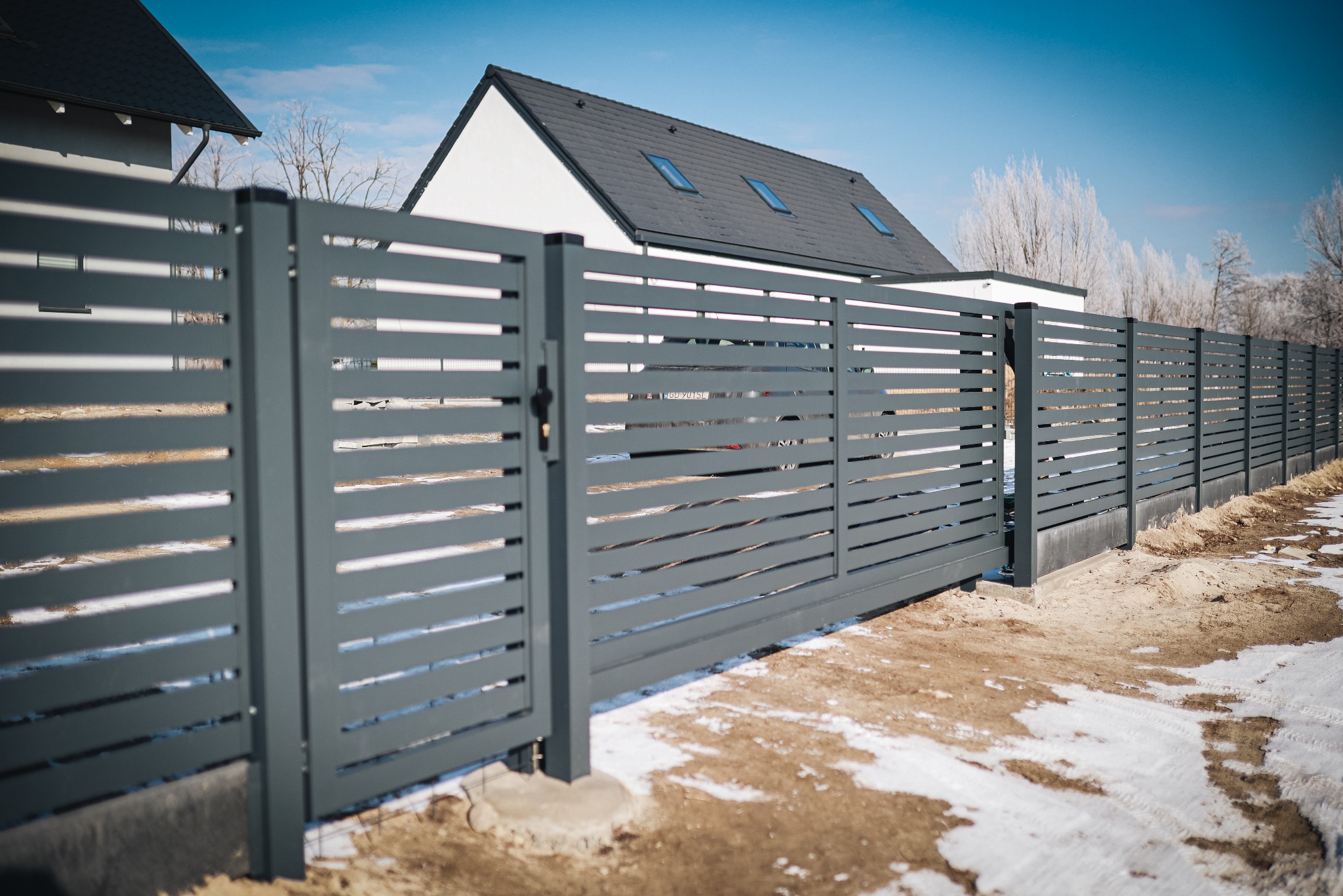 Nowoczesna, przesuwna brama wjazdowa z poziomych paneli w kolorze grafitowym, na tle domu z ciemną dachówką i błękitnego nieba. Zimowy krajobraz z resztkami śniegu.