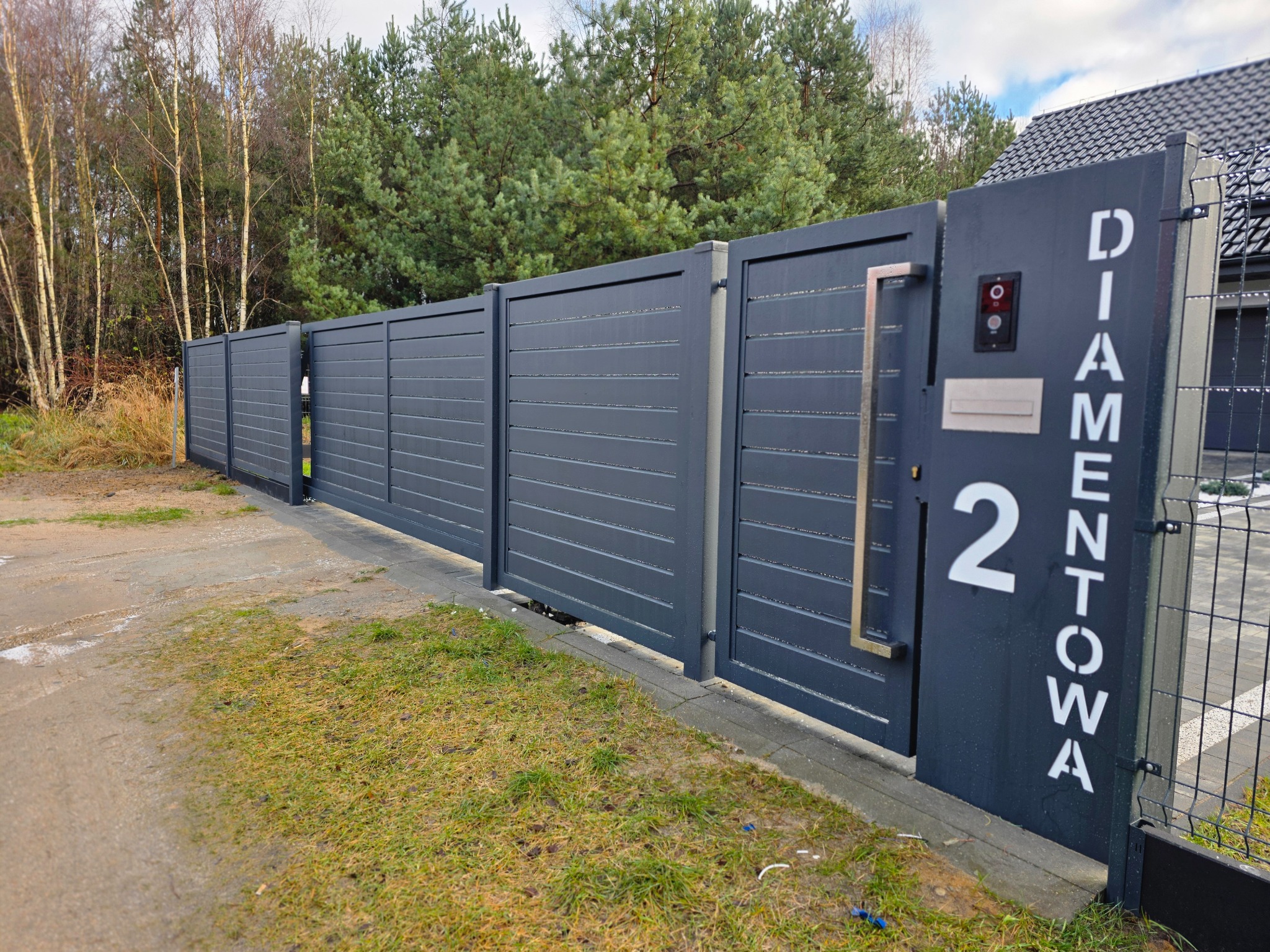 Nowoczesna, pozioma brama panelowa w kolorze grafitowym z numerem domu i domofonem, na tle zieleni i drzew. Minimalistyczny design, eleganckie wykończenie.