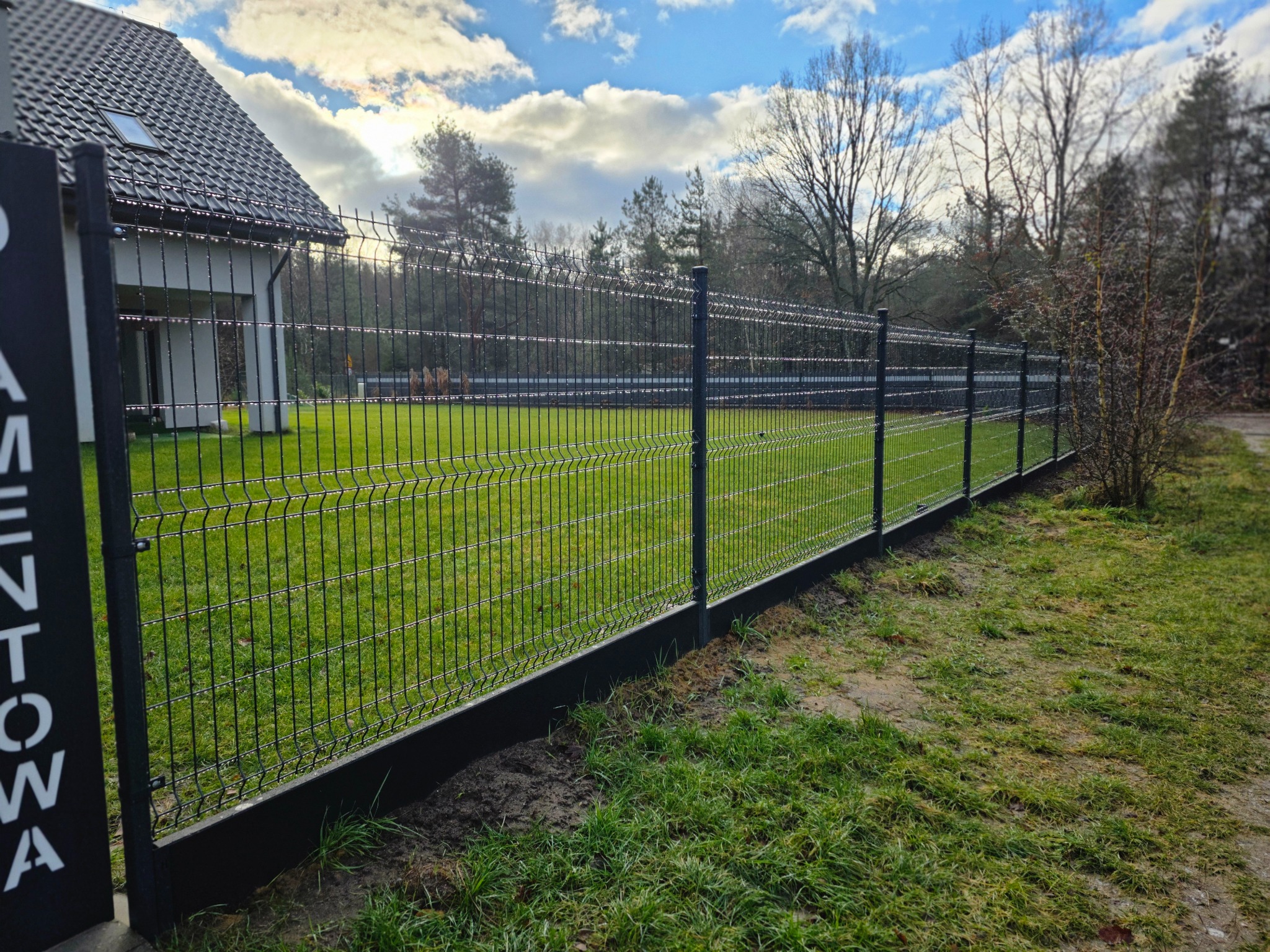 Nowoczesne ogrodzenie panelowe w kolorze grafitowym, z podmurówką, otaczające posesję z domem. Widok na równo przystrzyżony trawnik i błękitne niebo z chmurami.
