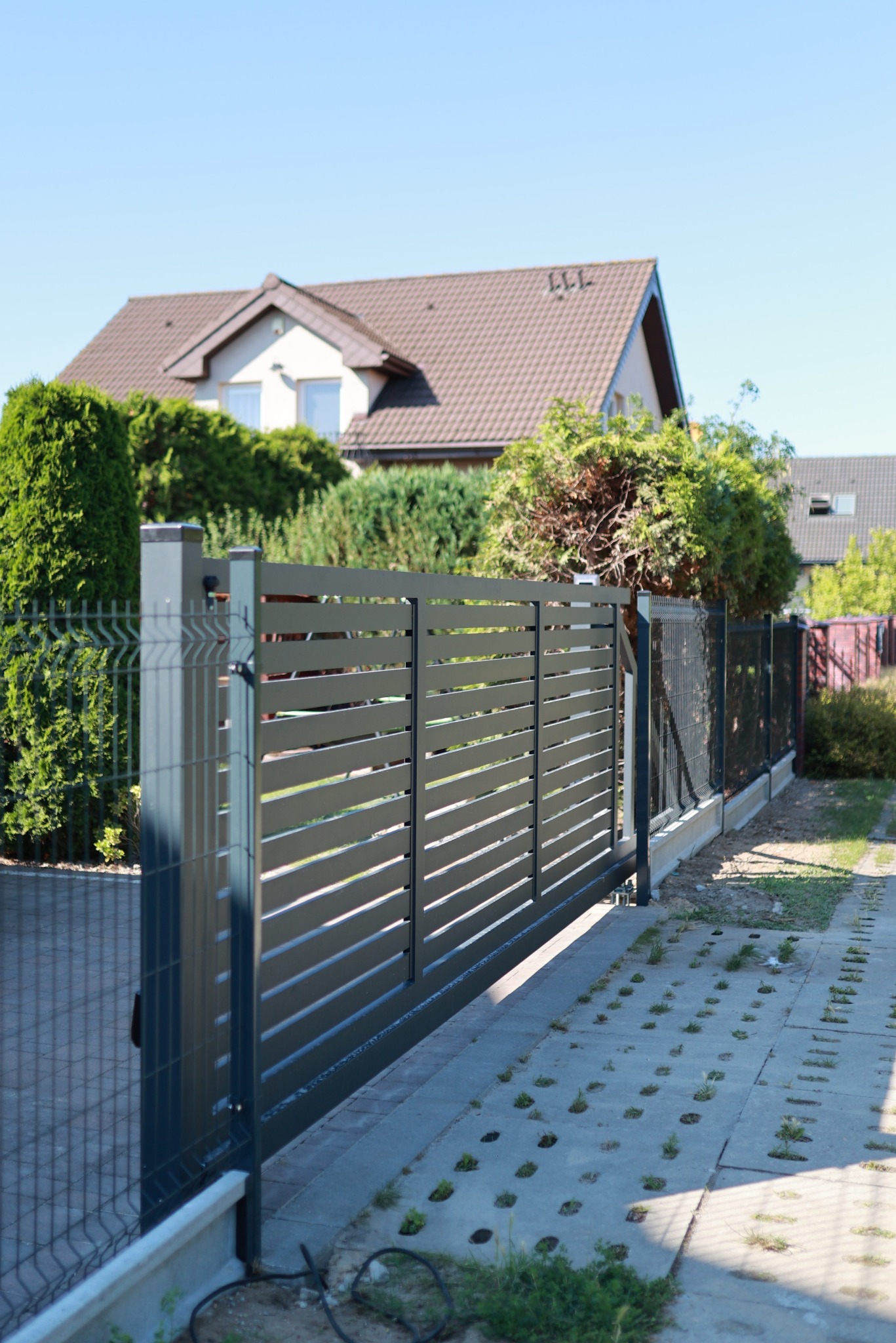 Nowoczesna, przesuwna brama wjazdowa z poziomymi panelami w kolorze grafitowym, z widocznym fragmentem siatki ogrodzeniowej i domu w tle.