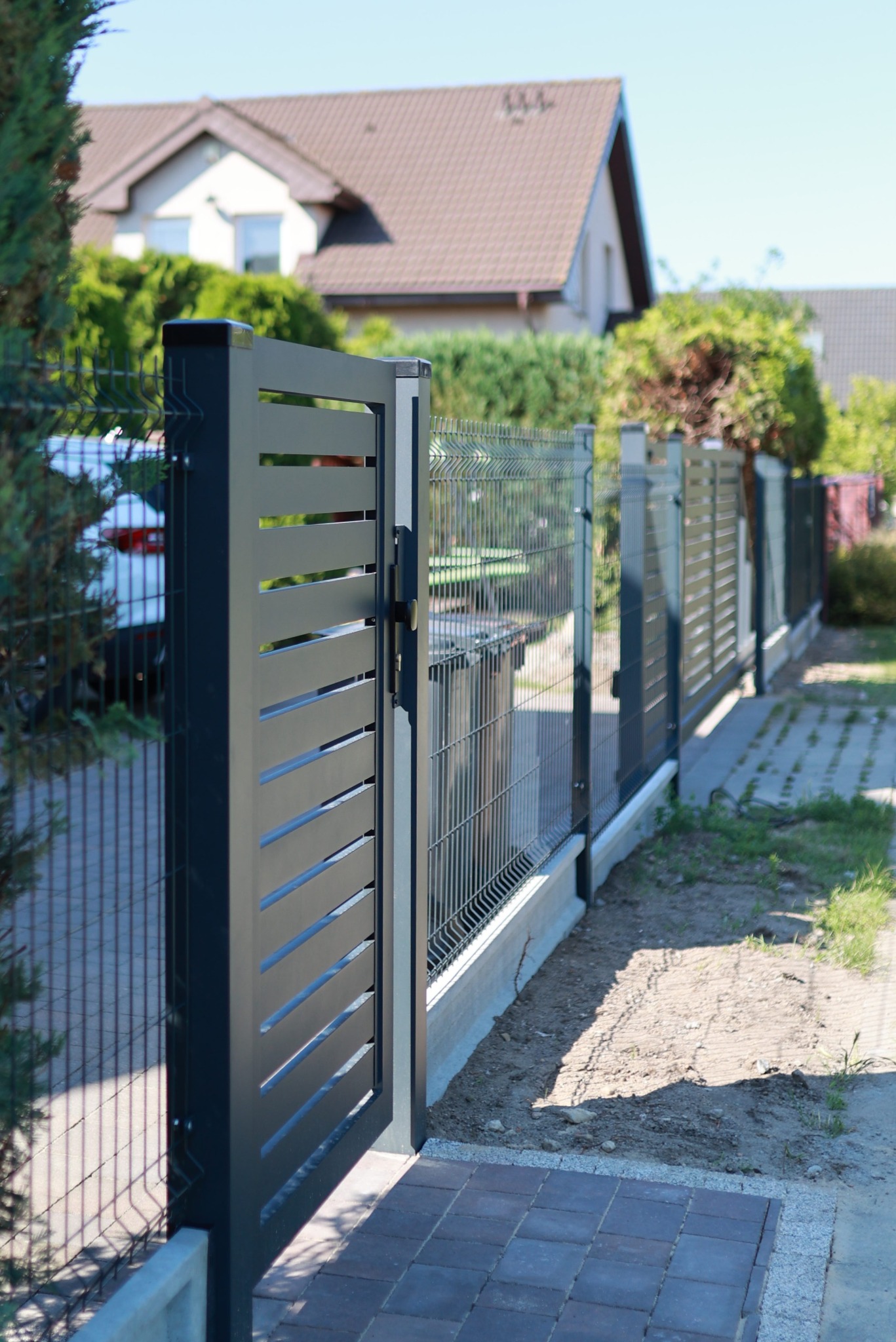 Nowoczesna brama wjazdowa z poziomymi szczeblami, połączona z ogrodzeniem panelowym. Widoczny fragment domu w tle. Estetyczne wykończenie.