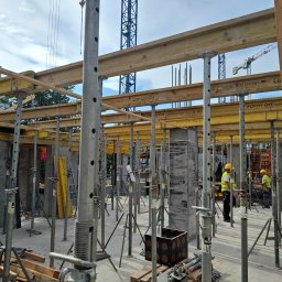 FIRMA OGÓLNOBUDOWLANA ANDRZEJ KIELIAN - Wznoszenie konstrukcji żelbetowej: szalunki stropowe na podporach, widoczni pracownicy w kaskach i kamizelkach odblaskowych. W tle żuraw budowlany i budynki.