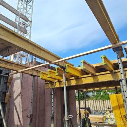 FIRMA OGÓLNOBUDOWLANA ANDRZEJ KIELIAN - Szczegółowy widok szalunków stropowych z żółtymi belkami i stalowymi podporami na budowie, z fragmentem żurawia w tle i pracownikiem w kasku.