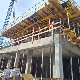 FIRMA OGÓLNOBUDOWLANA ANDRZEJ KIELIAN - Wznoszenie budynku żelbetowego: szalunki stropowe na podporach, widoczne zbrojenia i dźwigi w tle. Na dole worki z zaprawą Baumit MM100 i pracownik w kasku.