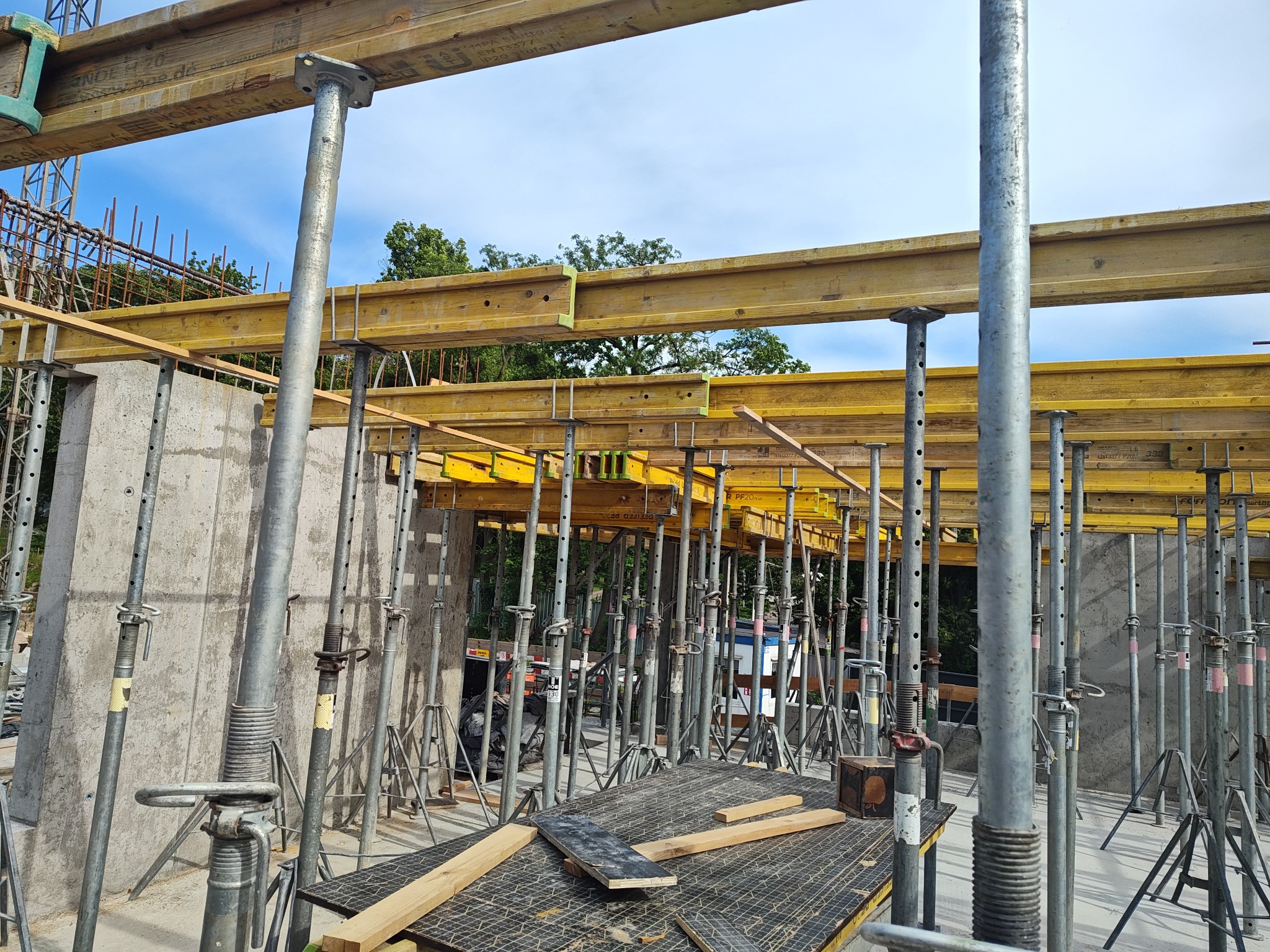 Szare ściany fundamentowe z żółtym szalunkiem i metalowymi podporami na tle błękitnego nieba. Widoczne zbrojenia i elementy konstrukcyjne.