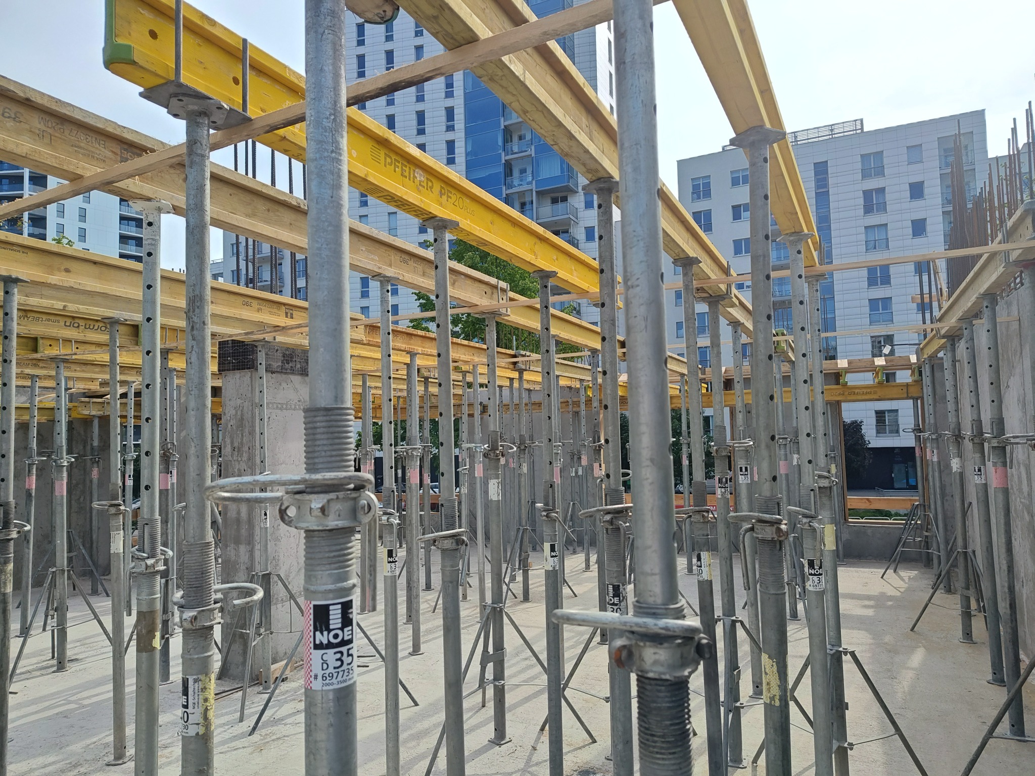 Szare podpory szalunkowe z żółtymi belkami na budowie, w tle budynki mieszkalne. Widoczne elementy konstrukcyjne i betonowe filary.