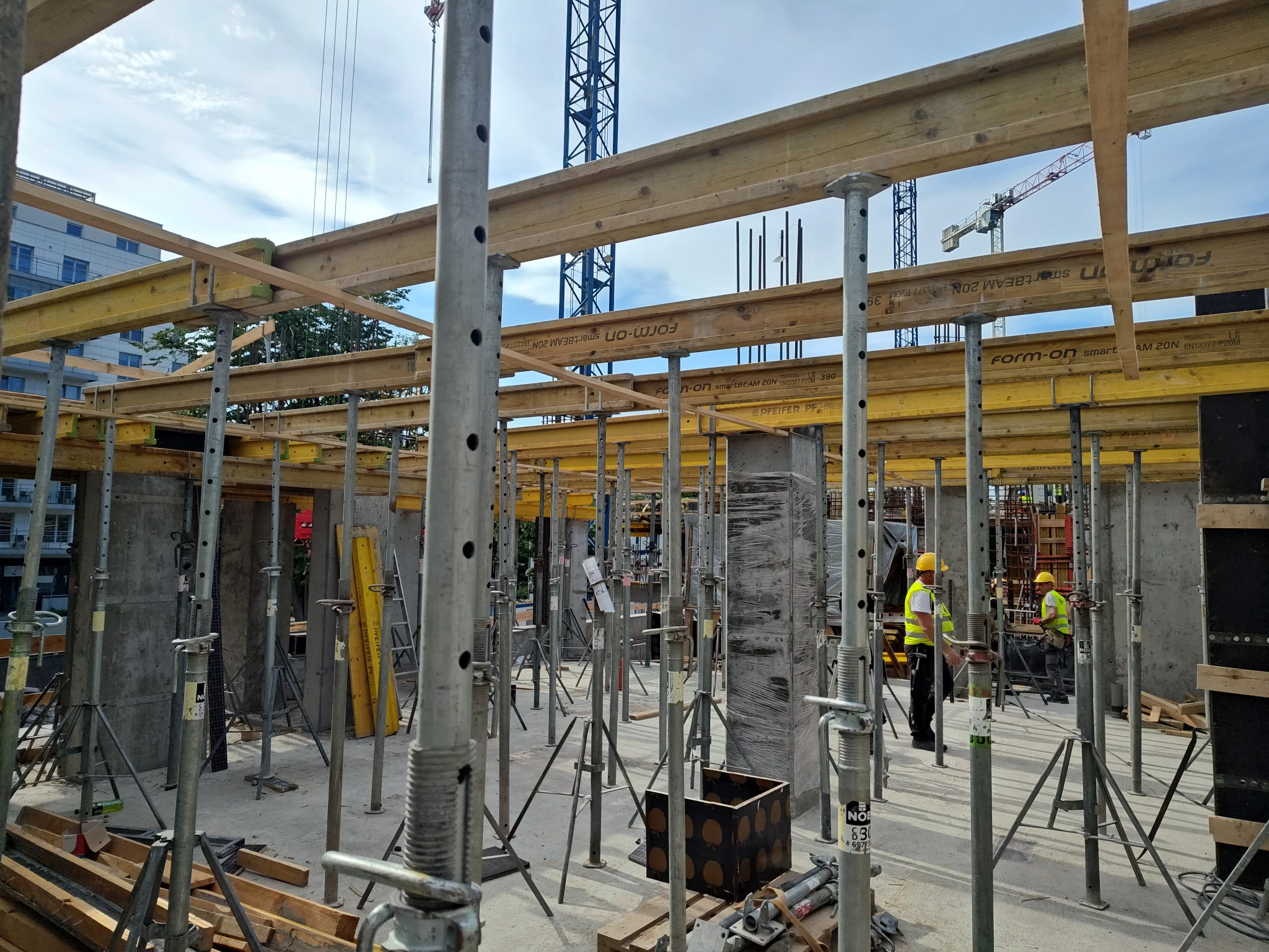 Wznoszenie konstrukcji żelbetowej: szalunki stropowe na podporach, widoczni pracownicy w kaskach i kamizelkach odblaskowych. W tle żuraw budowlany i budynki.