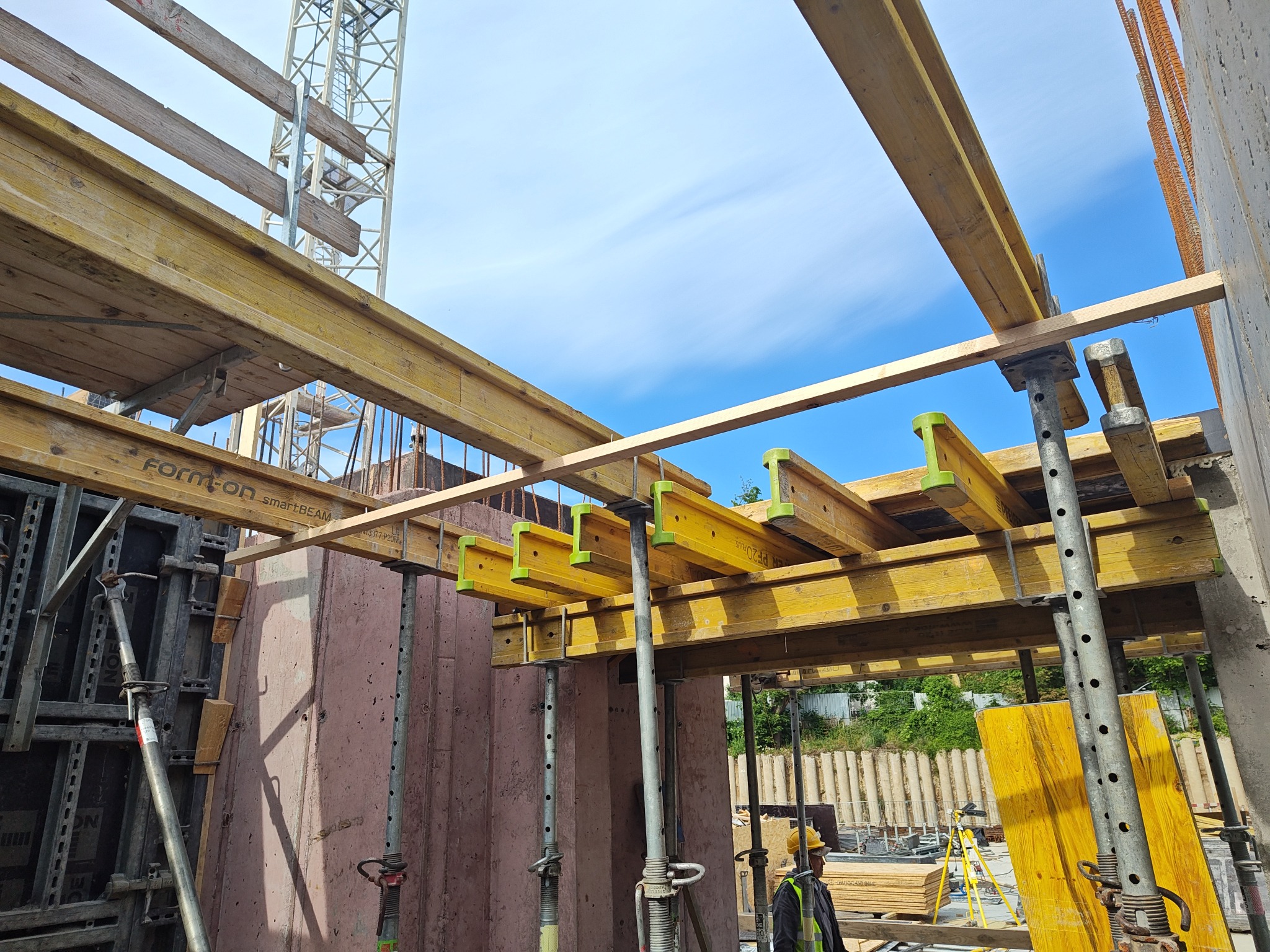 Szczegółowy widok szalunków stropowych z żółtymi belkami i stalowymi podporami na budowie, z fragmentem żurawia w tle i pracownikiem w kasku.