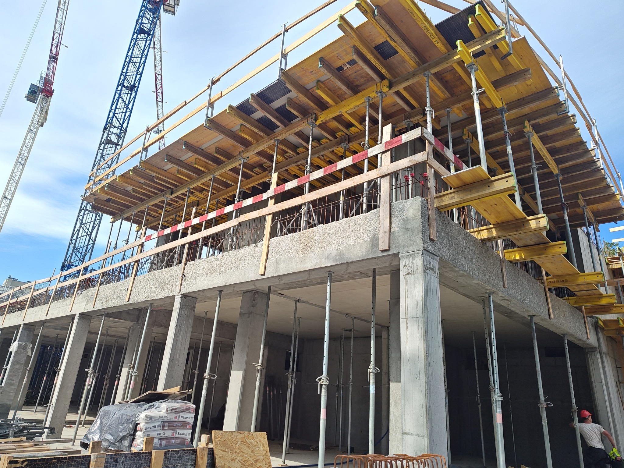 Wznoszenie budynku żelbetowego: szalunki stropowe na podporach, widoczne zbrojenia i dźwigi w tle. Na dole worki z zaprawą Baumit MM100 i pracownik w kasku.