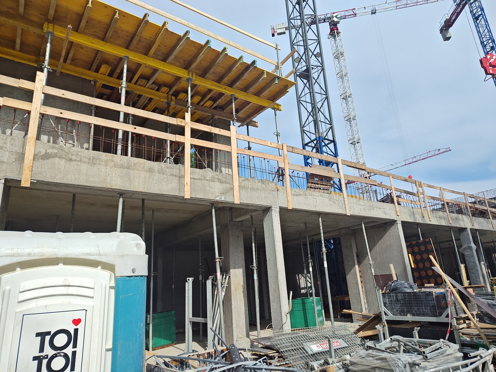Surowy szkielet budynku żelbetowego w trakcie budowy, widoczne szalunki stropowe, zbrojenia i żurawie w tle. Na pierwszym planie toaleta przenośna.
