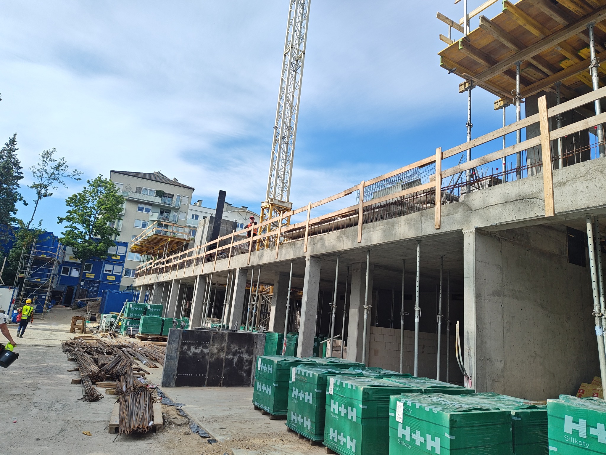 Budowa budynku żelbetowego w toku: widoczne zbrojenia, szalunki, kolumny wsporcze, palety z bloczkami H+H Silikaty i żuraw wieżowy na tle błękitnego nieba.