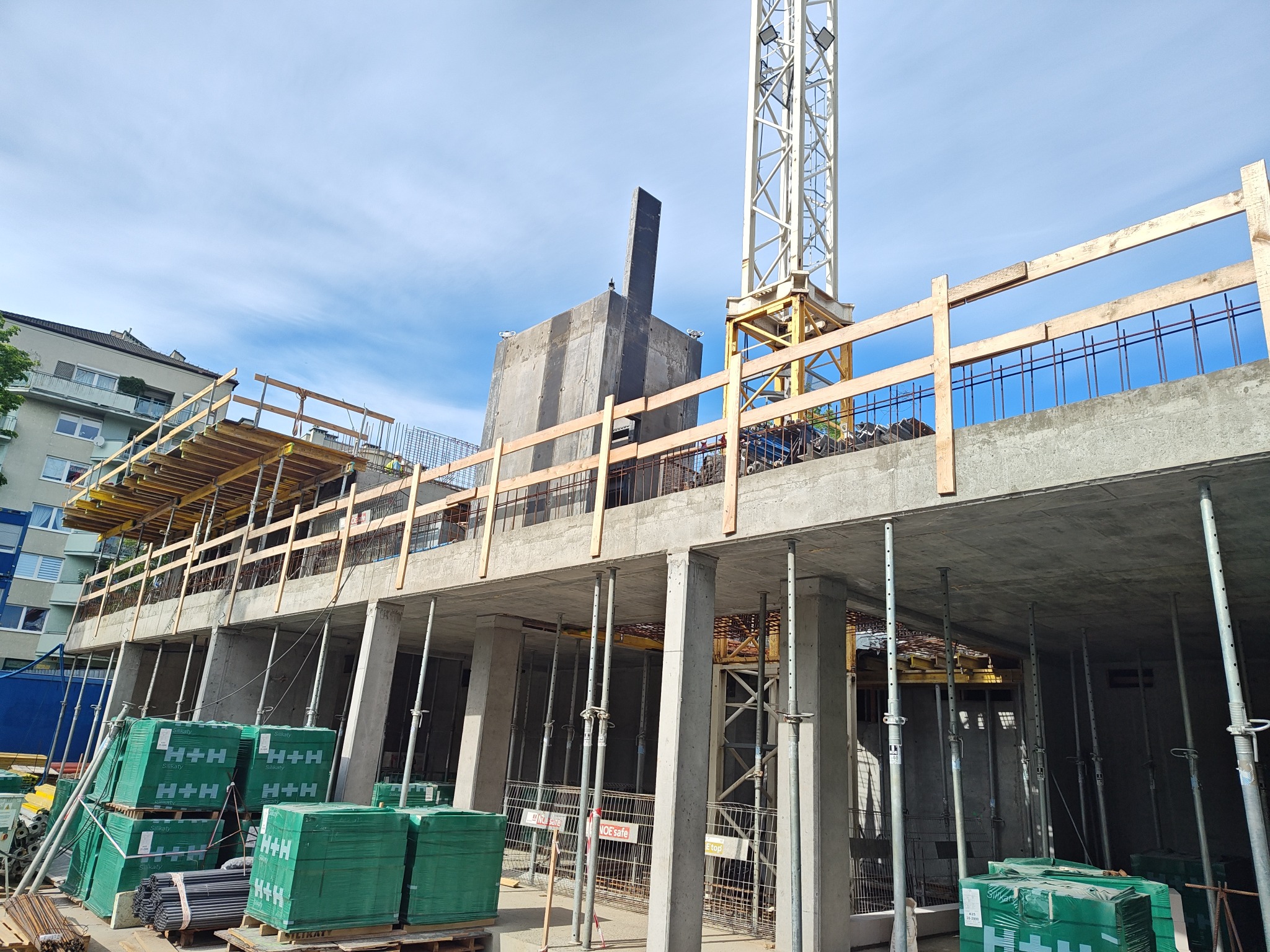 Budowa budynku żelbetowego w toku: widoczne zbrojenia, szalunki, podpory i dźwig. Na pierwszym planie palety z materiałami budowlanymi. Częściowo widoczne budynki mieszkalne w tle.