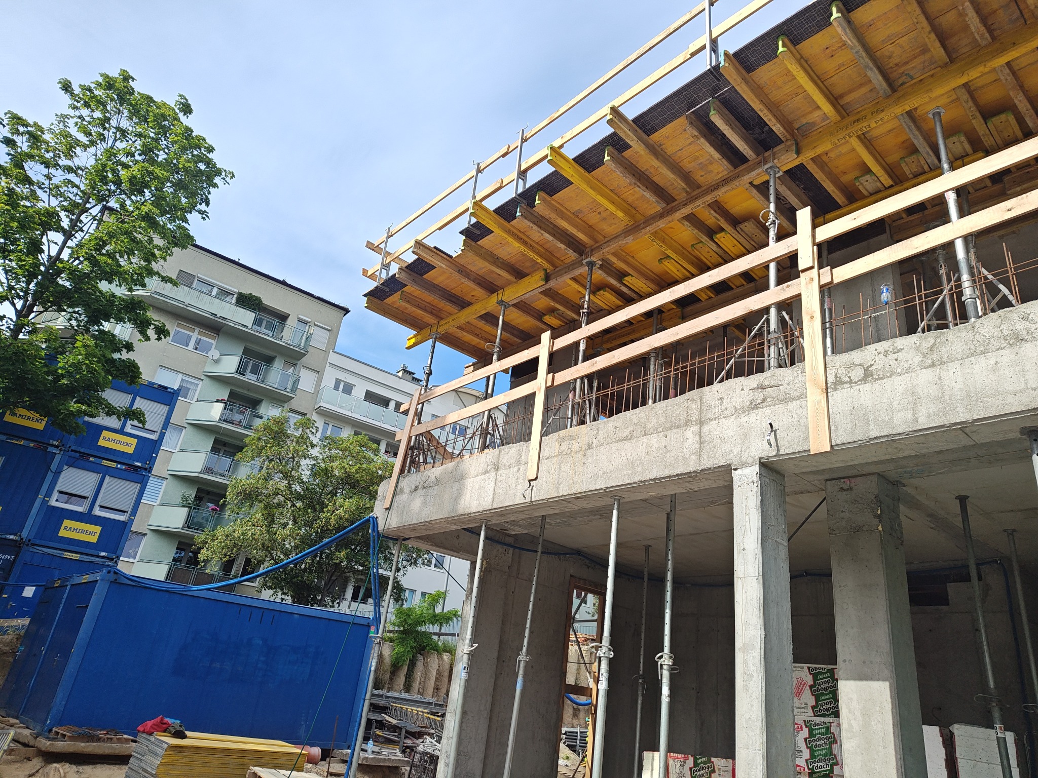 Budowa budynku żelbetowego: szalunek stropu na podporach, widoczne zbrojenie, kontenery budowlane w tle. Ujęcie z dołu, w tle budynek mieszkalny i drzewa.