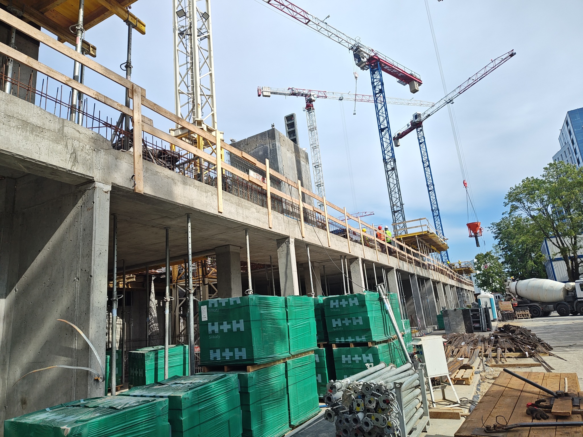 Budowa budynku żelbetowego w toku: widoczne szalunki, zbrojenia, dźwigi i materiały budowlane H+H. Betonowe elementy konstrukcyjne w surowym stanie.
